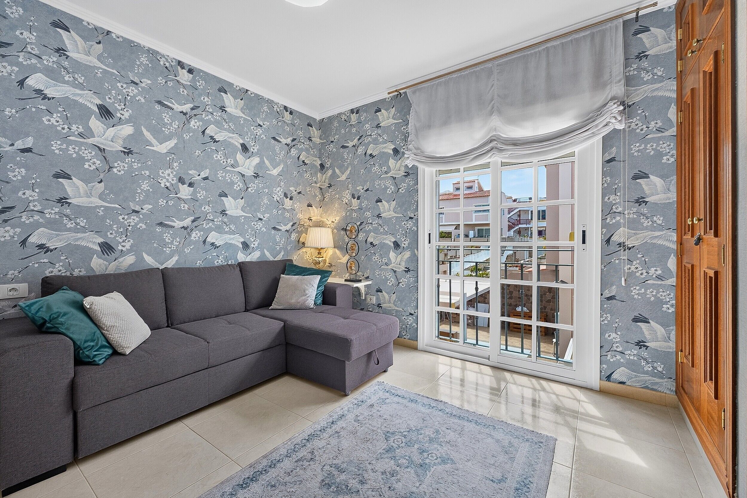 A living room with a grey sectional sofa, a patterned wallpaper, a large window with a view of buildings, and a wooden door.