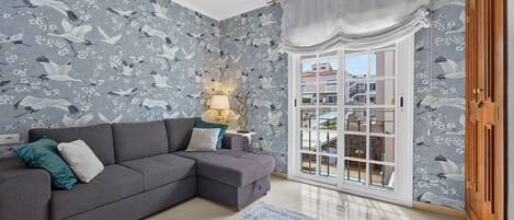 A living room with a grey sectional sofa, a patterned wallpaper, a large window with a view of buildings, and a wooden door.