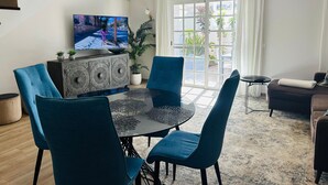 Een moderne woonkamer met een eettafel, stoelen, een televisietoestel en een groot raam met gordijnen.