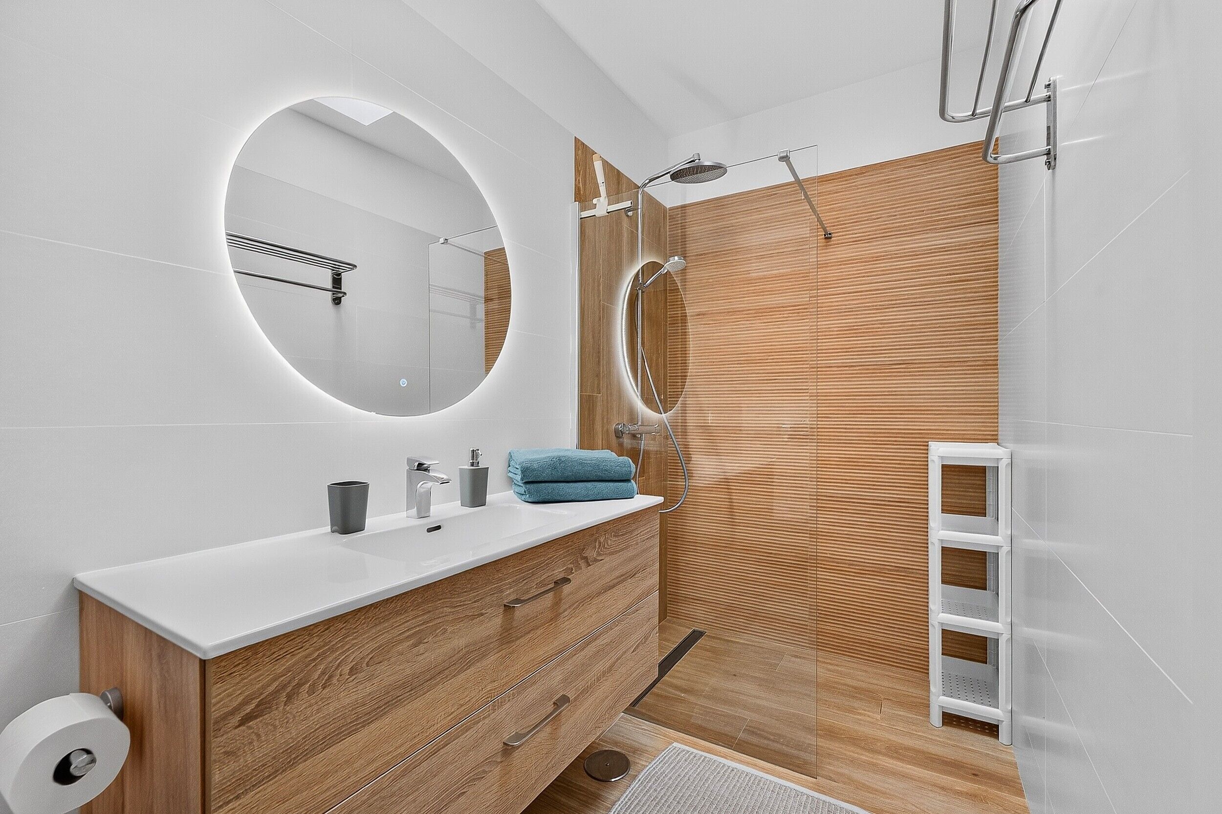 Un baño moderno con tocador de madera, espejo redondo, ducha y lavamanos blanco.