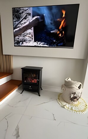 A room with a television mounted on the wall displaying a fire scene, a small black wood-burning stove, and a white ceramic pot on a woven mat.