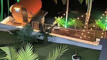 A modern outdoor structure with a curved roof, surrounded by tropical vegetation and illuminated by string lights.