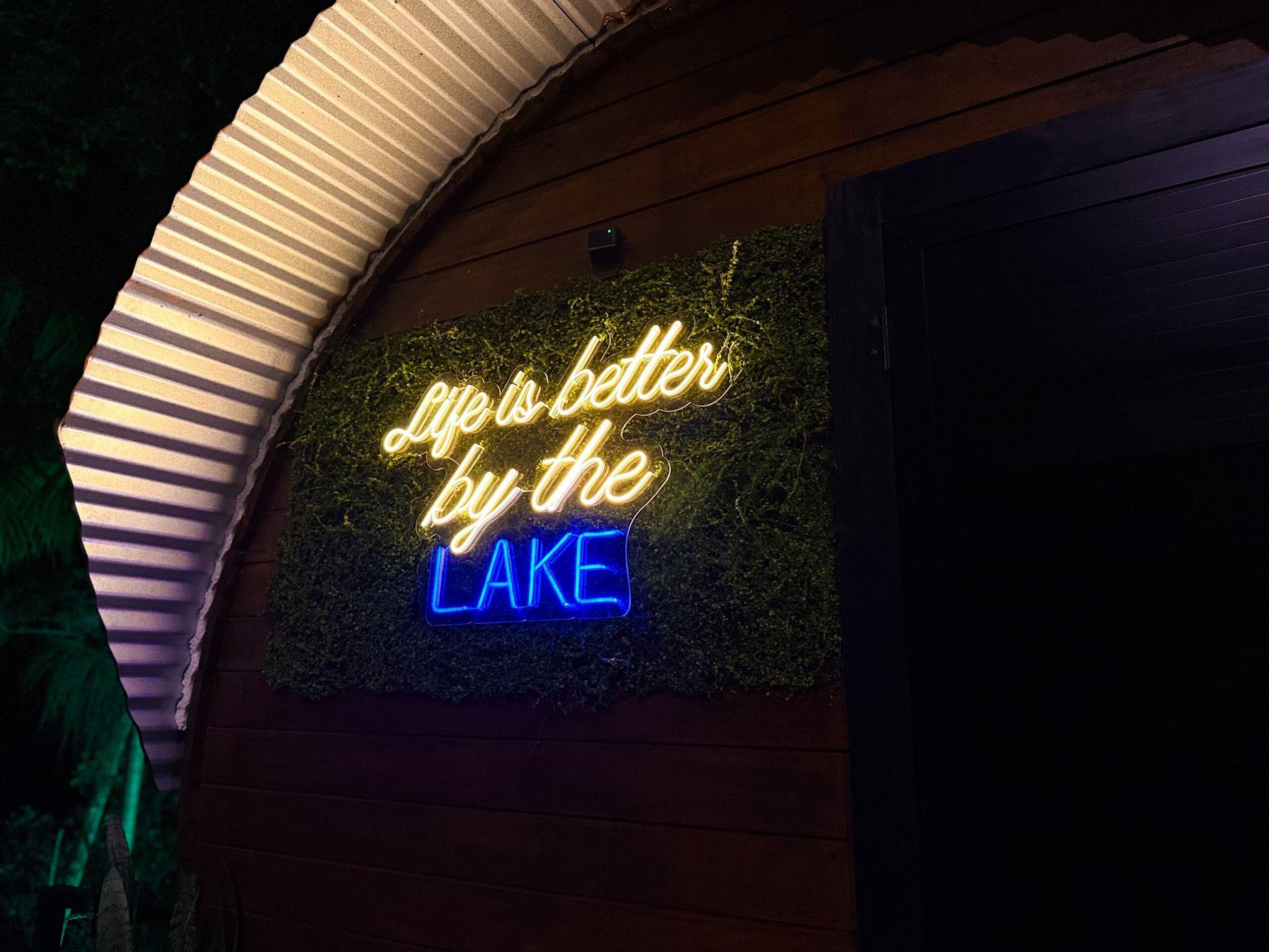 Un letrero de neón con el texto 'Life is better by the lake' frente a una pared con textura.