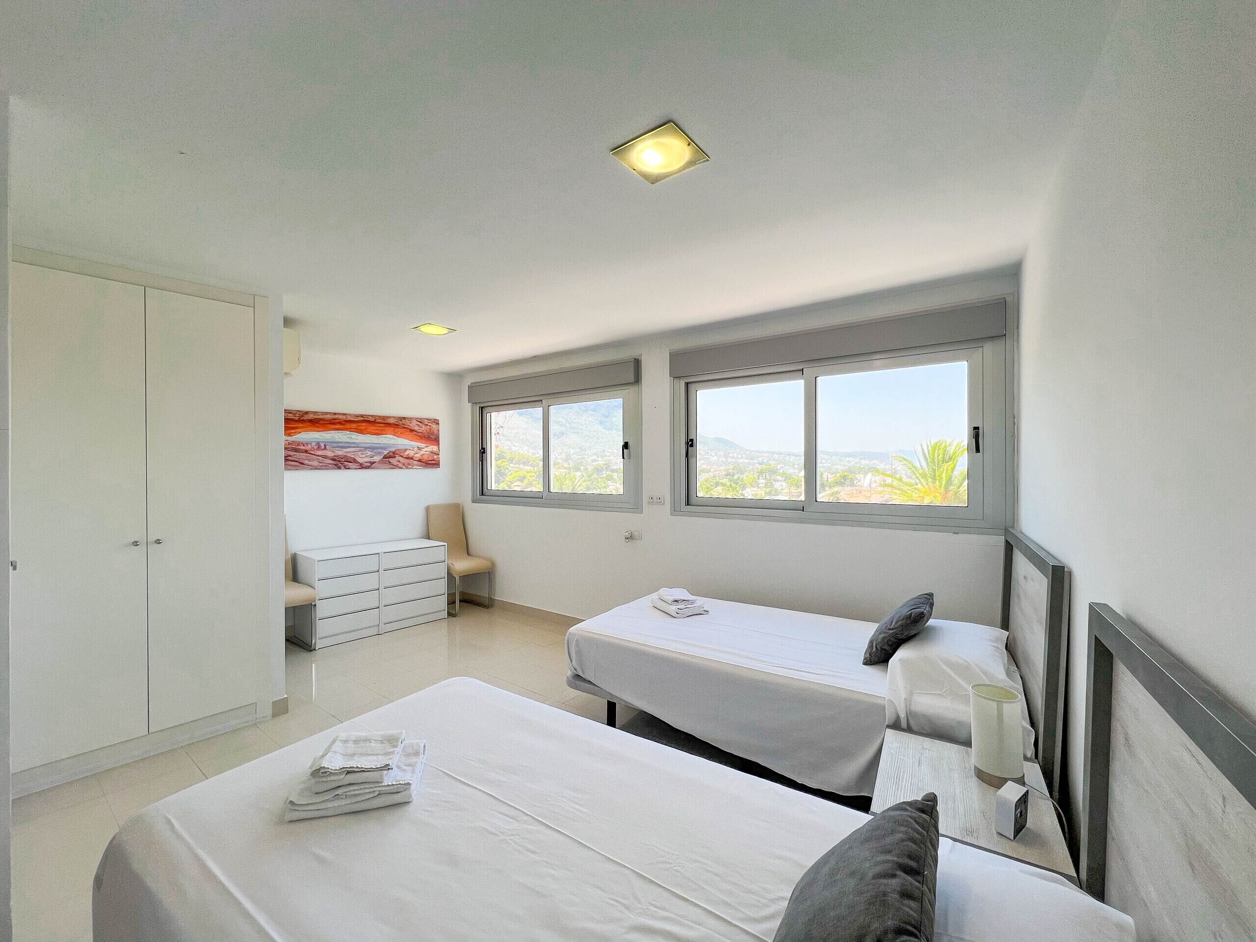 A bedroom with two beds, a wardrobe, a chair, and a window with a city view.
