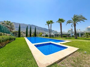 Ein rechteckiger Swimmingpool mit glasklarer blauer Wasseroberfläche, umgeben von einer gepflegten Rasenfläche und Palmen, mit einem modernen Gebäudekomplex im Hintergrund.