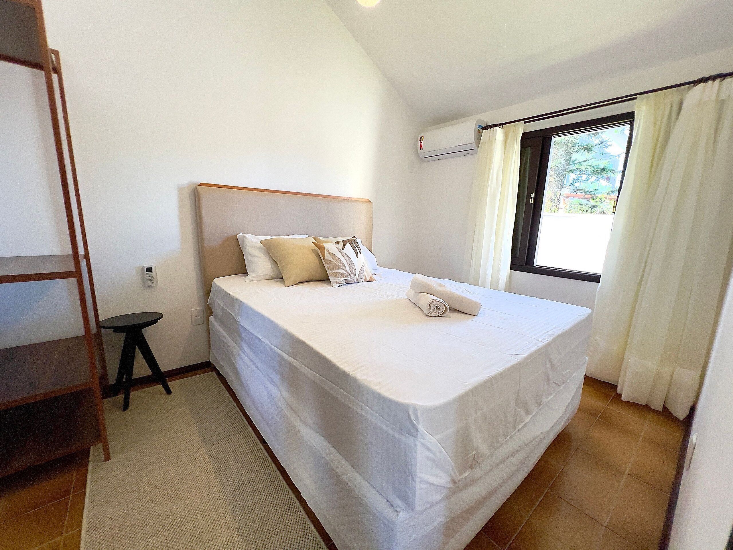 Un dormitorio con una cama, una mesita, aire acondicionado instalado en la pared y una ventana con cortinas.
