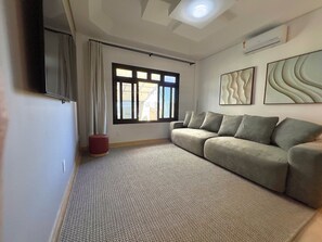 Una sala de estar con sofá, ventana con cortinas, un reposapiés rojo y arte abstracto en la pared.