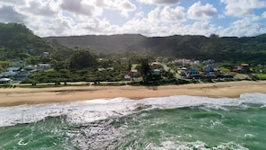 A coastal view with a beach, ocean waves, and a town with buildings and greenery in the background.