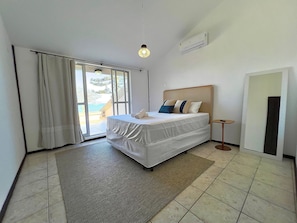 A hotel room with a bed, a small bedside table, a view through a sliding door, and a ceiling-mounted air conditioning unit.