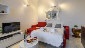 A modern hotel room with a flat-screen TV, a bed with red and grey bedding, a white console table with a bottle and glasses, and a unique wall mirror. - Lodi, 65 – Your Milan Base (Milan)