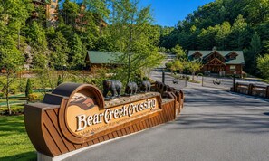 Indicador de fusta de Bear Creek Crossing amb una escultura d'ós, envoltat d'arbres i un edifici al fons.