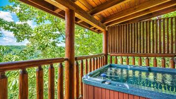 A wooden deck with a hot tub, surrounded by lush greenery and a clear sky.