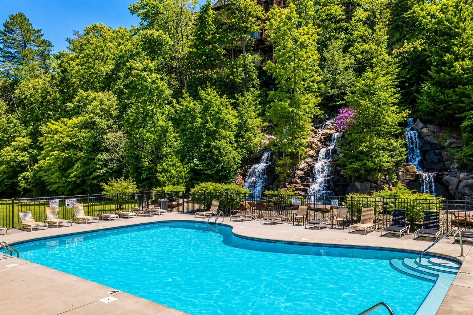 A swimming pool with multiple waterfalls, surrounded by lush green trees and a clear sky.