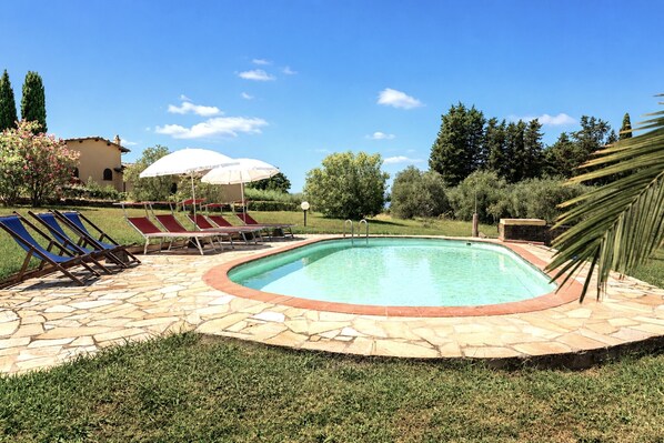 Ein Swimmingpool mit Steinrand, umgeben von Gras und Bäumen, mit Liegestühlen und Sonnenschirmen.
