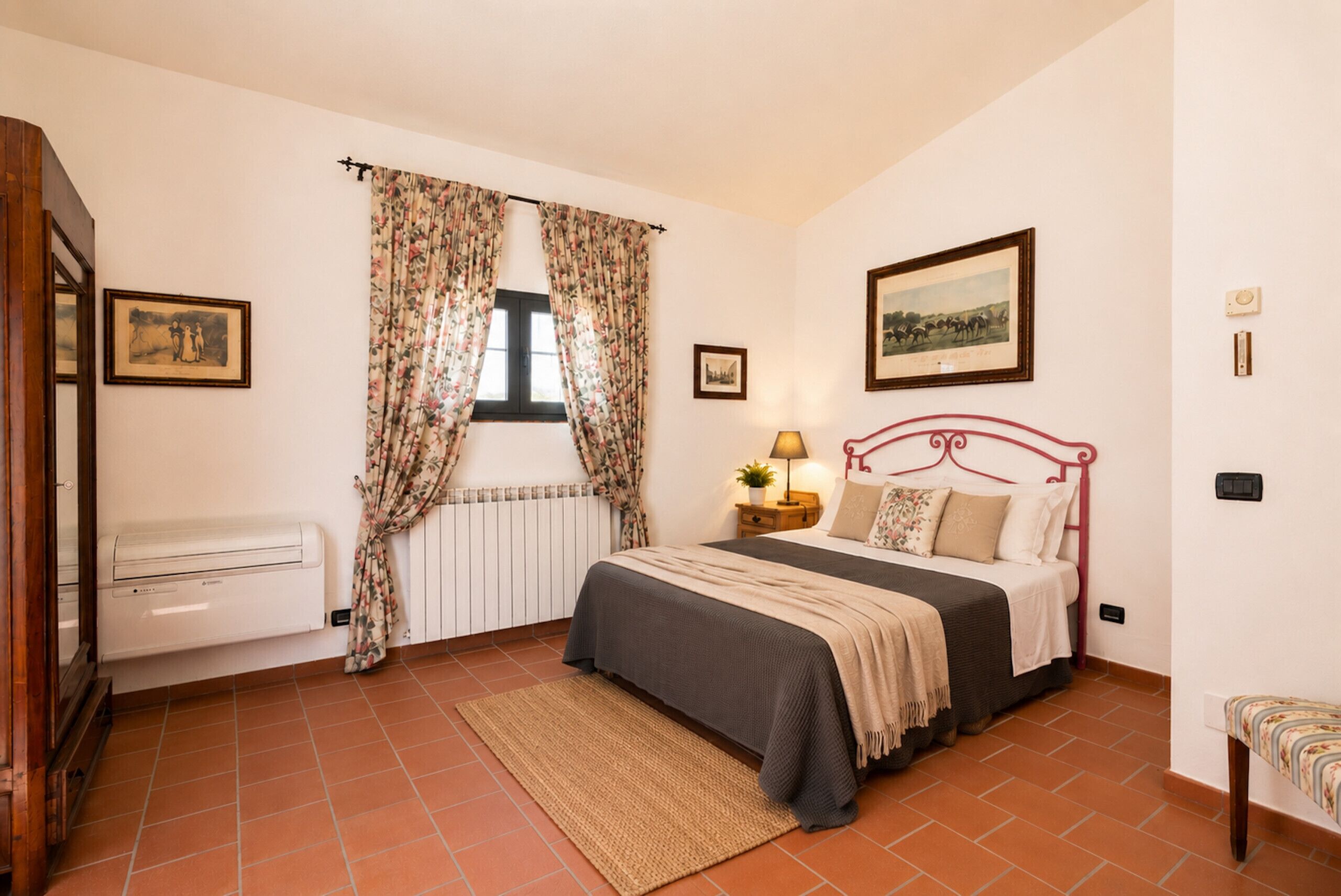 A bedroom with a bed, a radiator, a window with curtains, a bedside table with a lamp, and a picture on the wall.