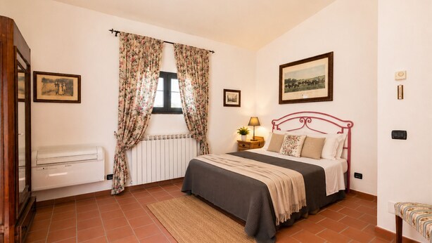 A bedroom with a bed, a radiator, a window with curtains, a bedside table with a lamp, and a framed picture on the wall. - Gaetano Spadaro Estate, La Rosa Room (Capannori)