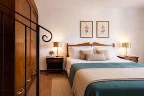 Ein ordentlich eingerichtetes Schlafzimmer mit einem Holzschrank, einem Bett mit grüner Bettdecke, zwei Nachttischlampen und zwei gerahmten Pflanzenbildern an der Wand.