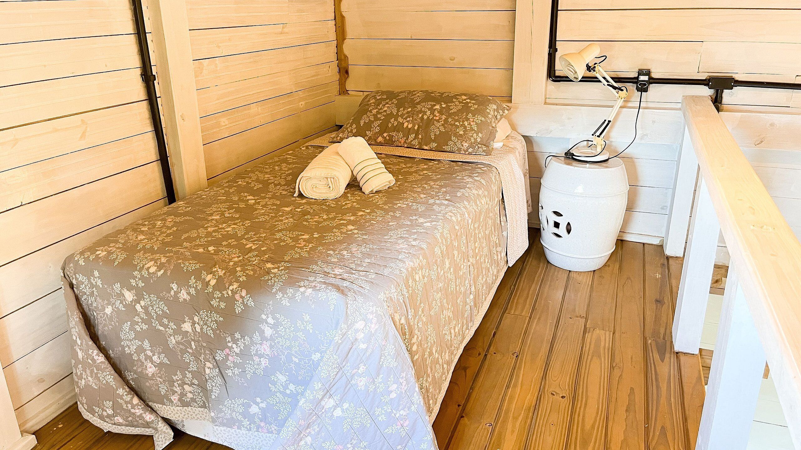 A single bed with a floral bedspread and a rolled towel, a white trash bin, and a wooden bench.