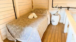 A single bed with a floral bedspread and a rolled towel, a white trash bin, and a wooden bench.