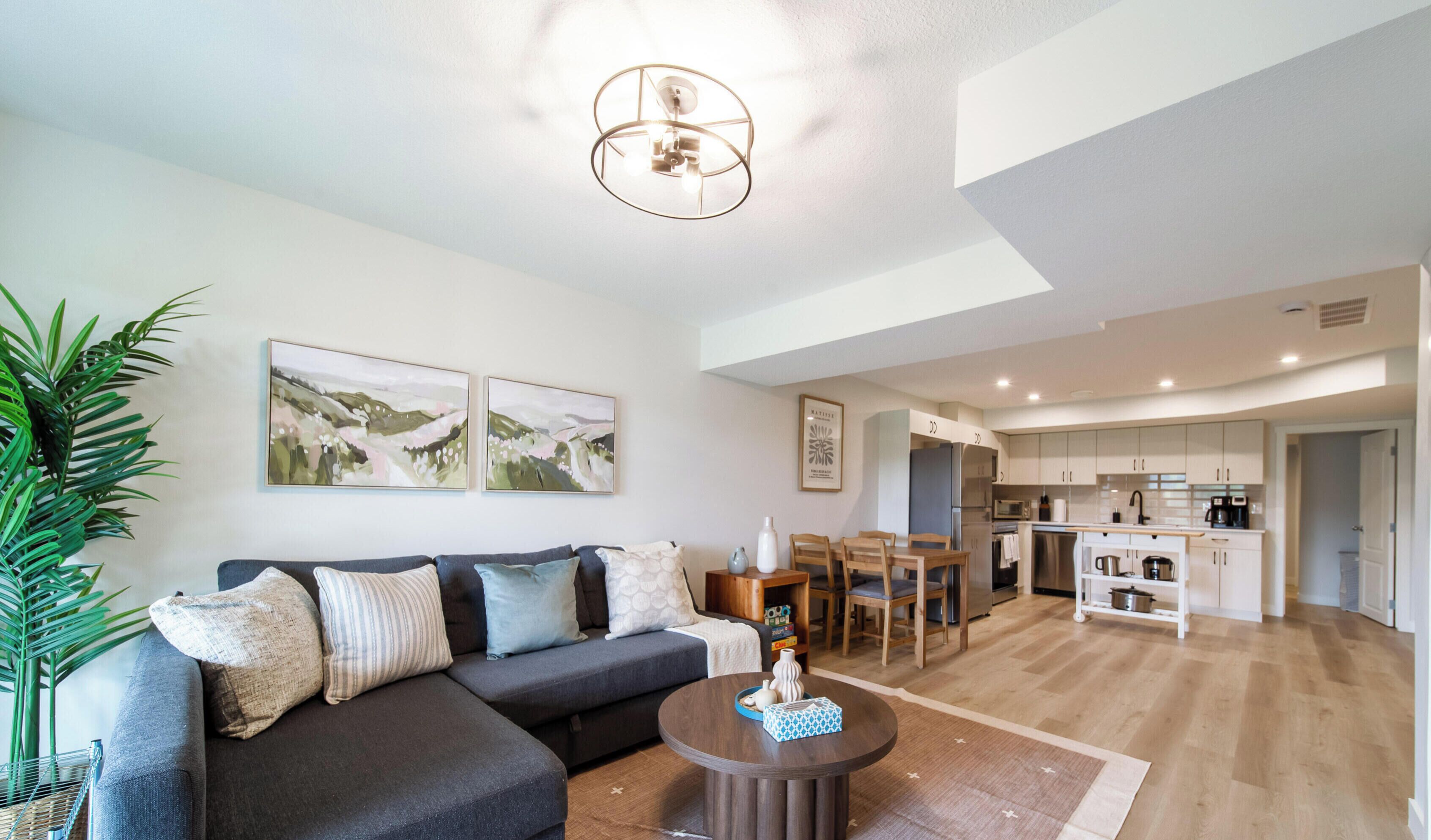 Un salon moderne avec un canapé d’angle, un coin repas avec une table et des chaises en bois, et une cuisine avec des meubles blancs.