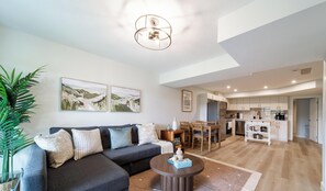 Un salon moderne avec un canapé d’angle, un coin repas avec une table et des chaises en bois, et une cuisine avec des meubles blancs.