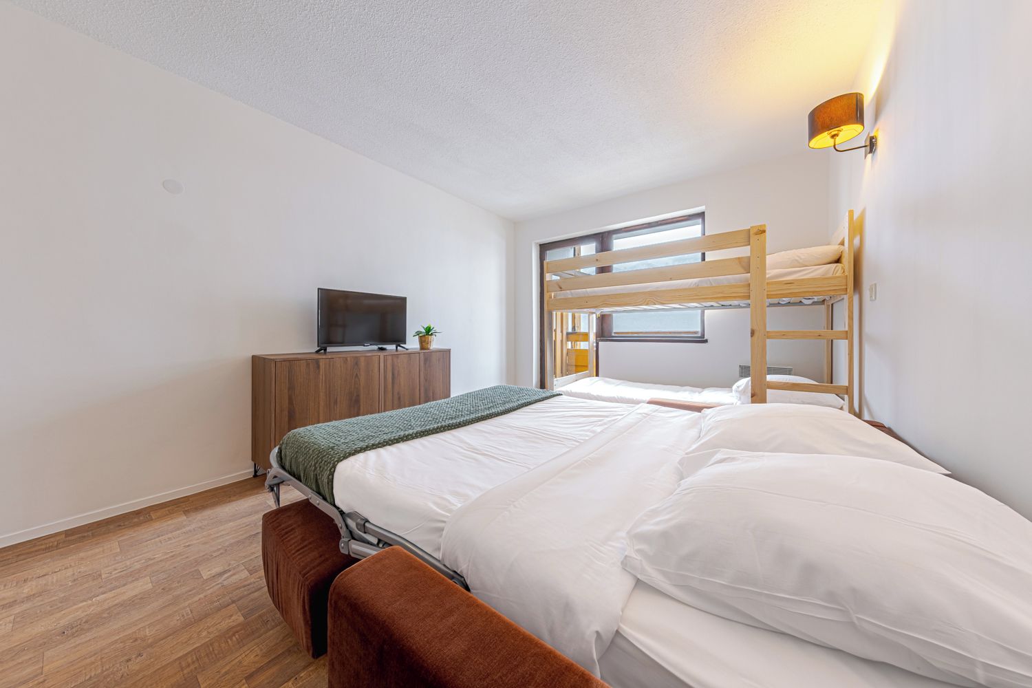 Une chambre avec un lit superposé, un téléviseur posé sur un meuble en bois et une fenêtre laissant entrer la lumière naturelle.
