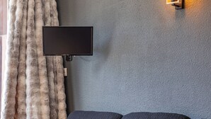 Ein modernes Wohnzimmer mit grauer Wand mit Struktur, einem fest montierten Flachbildfernseher, einem dunkelgrauen Sofa, einem Couchtisch aus Holz mit einem Topf und Pflanze und einem großen Fenster mit weißem Vorhang.