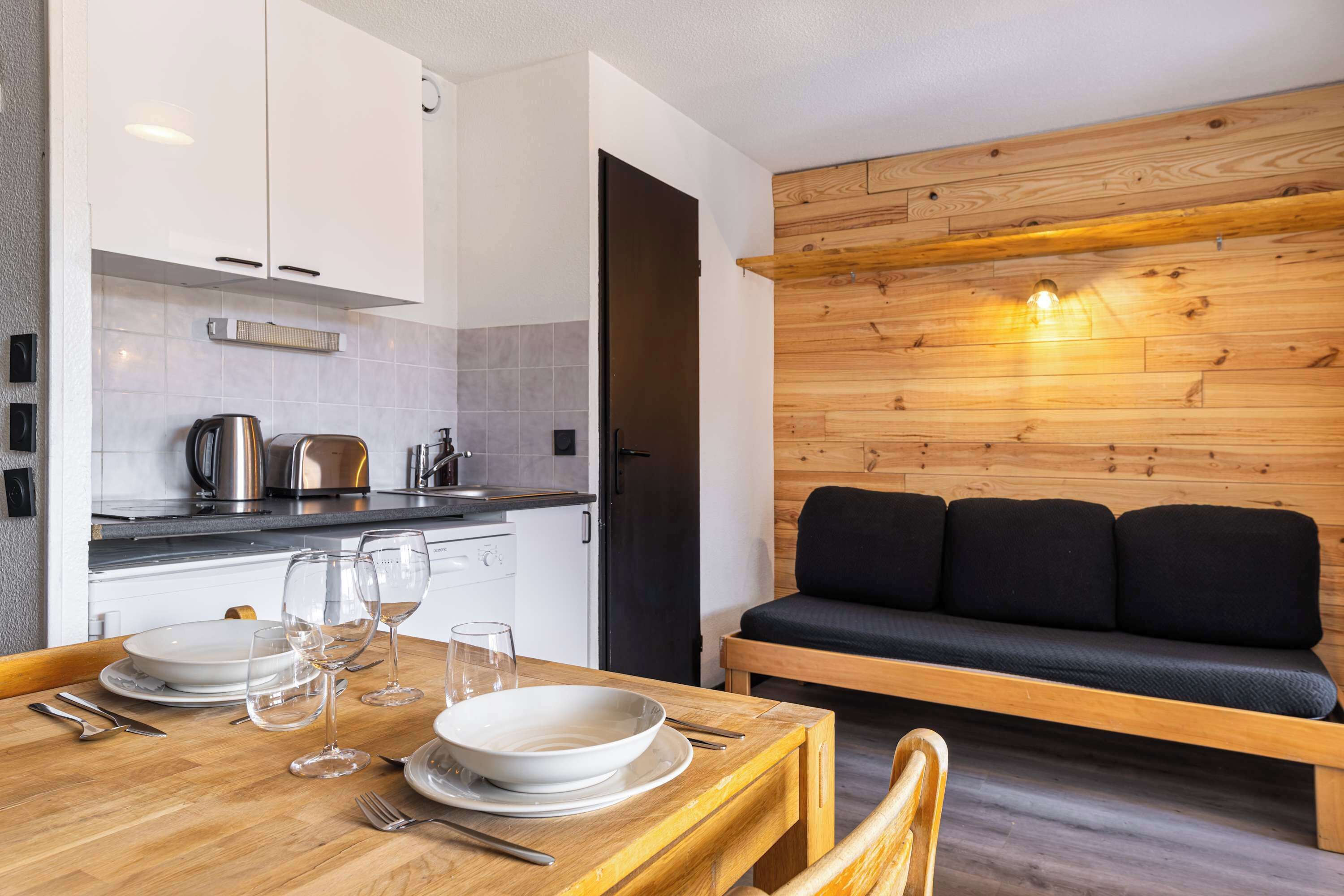 Une cuisine moderne avec des meubles blancs, une table à manger en bois mise avec des assiettes et des verres, et un canapé noir placé contre un mur en bois.