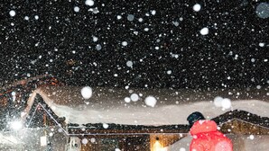 Eine Person ist auf einem verschneiten Hang beim Skifahren, im Hintergrund ist ein Gebäude zu sehen.