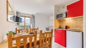 Ein Essbereich mit Holzkästen und einem Tisch für zwei Personen, eine Küche mit roten Schränken und einer Mikrowelle sowie ein Fenster mit Blick auf Grünflächen.
