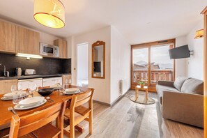 A modern kitchen with a microwave, toaster, and coffee maker. There is a dining table set for four with wine glasses and plates. A sofa and a TV are visible in the living area. - Residence Altineige, Ski In-Out with WiFi in Val Thorens, “V9” by Versant Collection (Val Thorens)