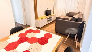 Een moderne woonkamer met een bank, een salontafel met een geometrisch vloerkleed, een televisie op een dressoir en een raam met gordijnen.