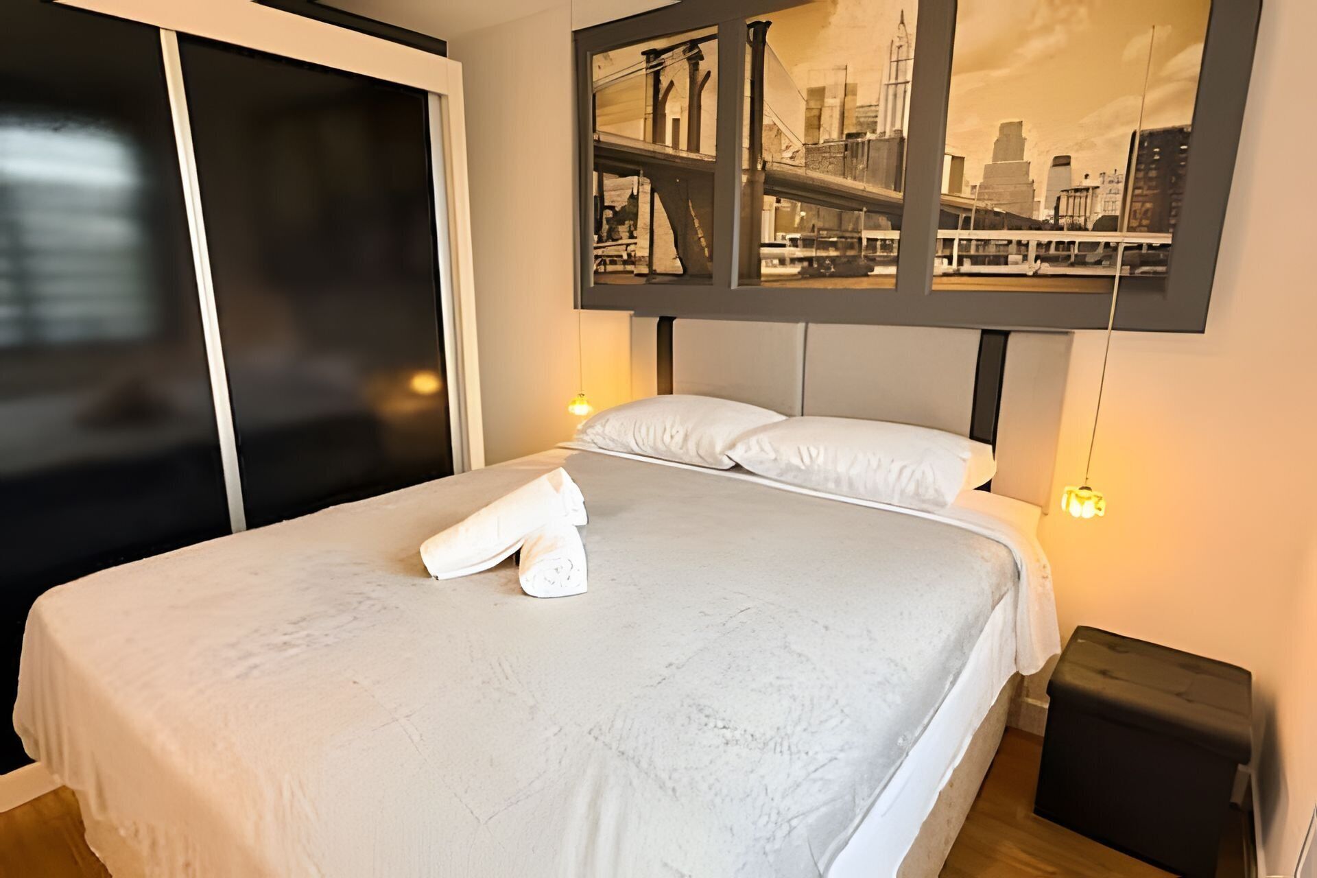 Habitación de hotel con una cama, una mesita de noche y una obra de arte en la pared que muestra un paisaje urbano.
