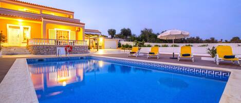 A swimming pool with a clear blue water surface, surrounded by lounge chairs and a building with a tiled roof.