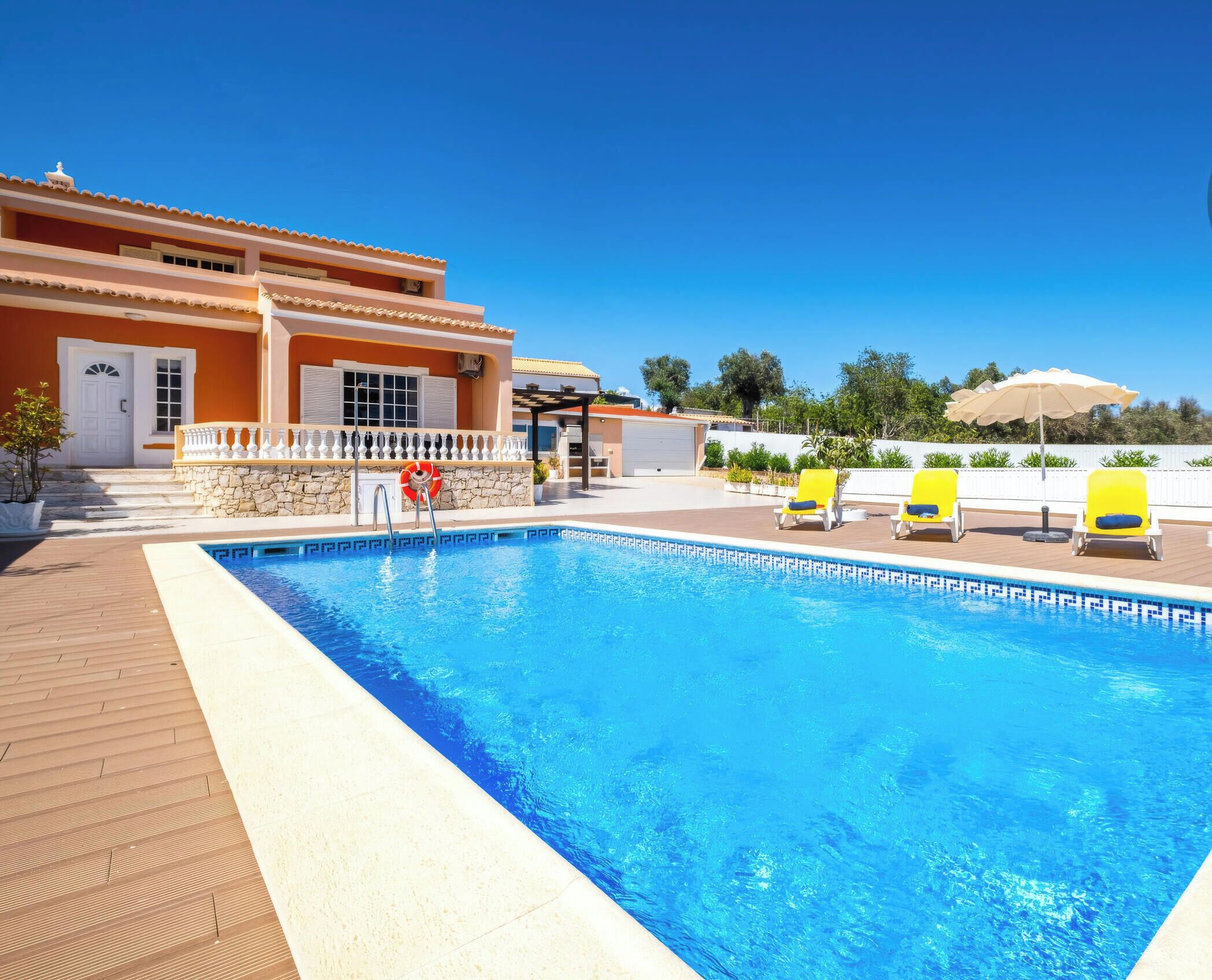 Une piscine avec une terrasse en bois, un espace couvert agrémenté d’une pergola et un bâtiment doté d’une porte et de fenêtres blanches.