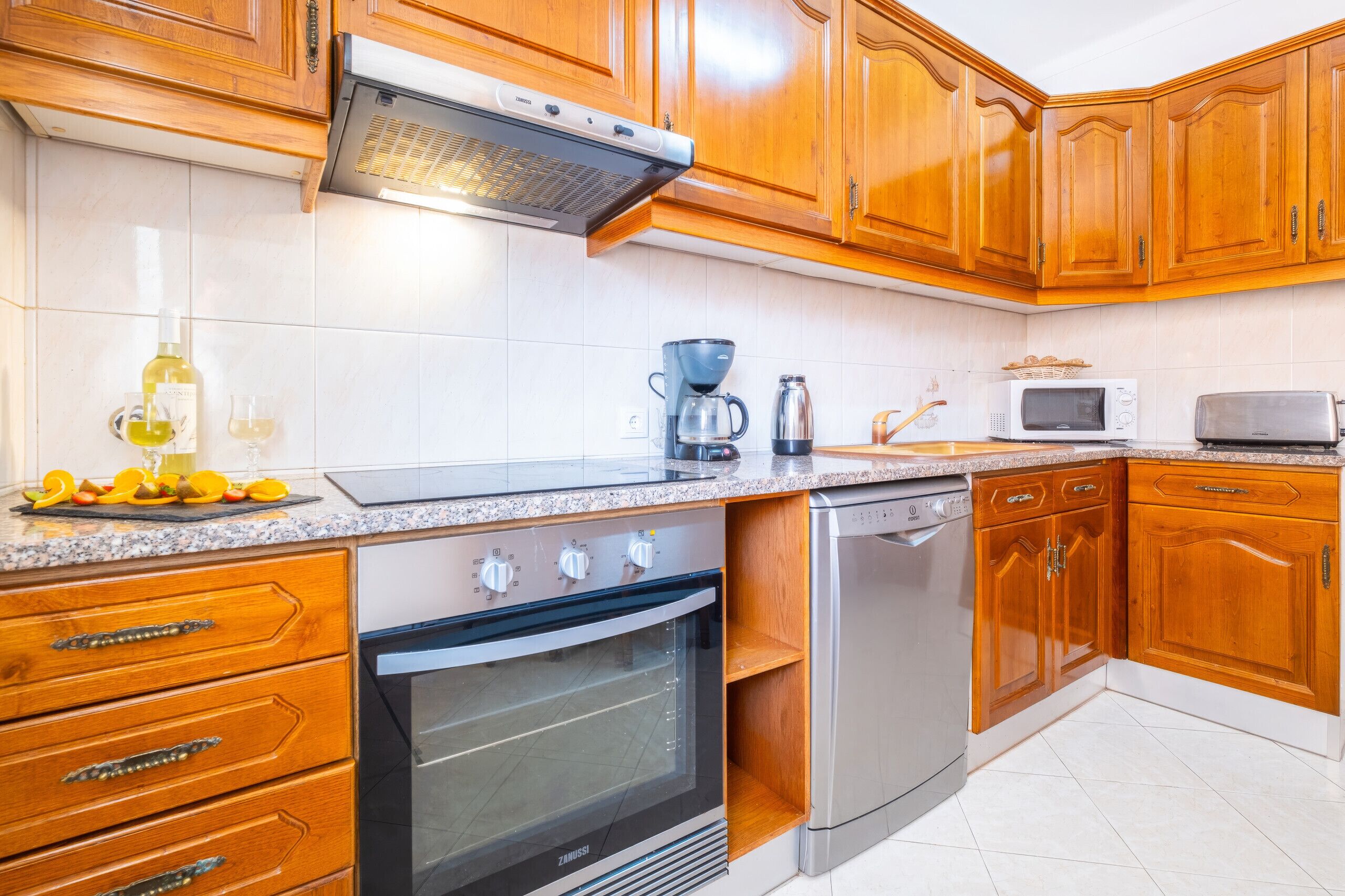 Una cocina con armarios de madera, horno empotrado, lavavajillas, cafetera y microondas.