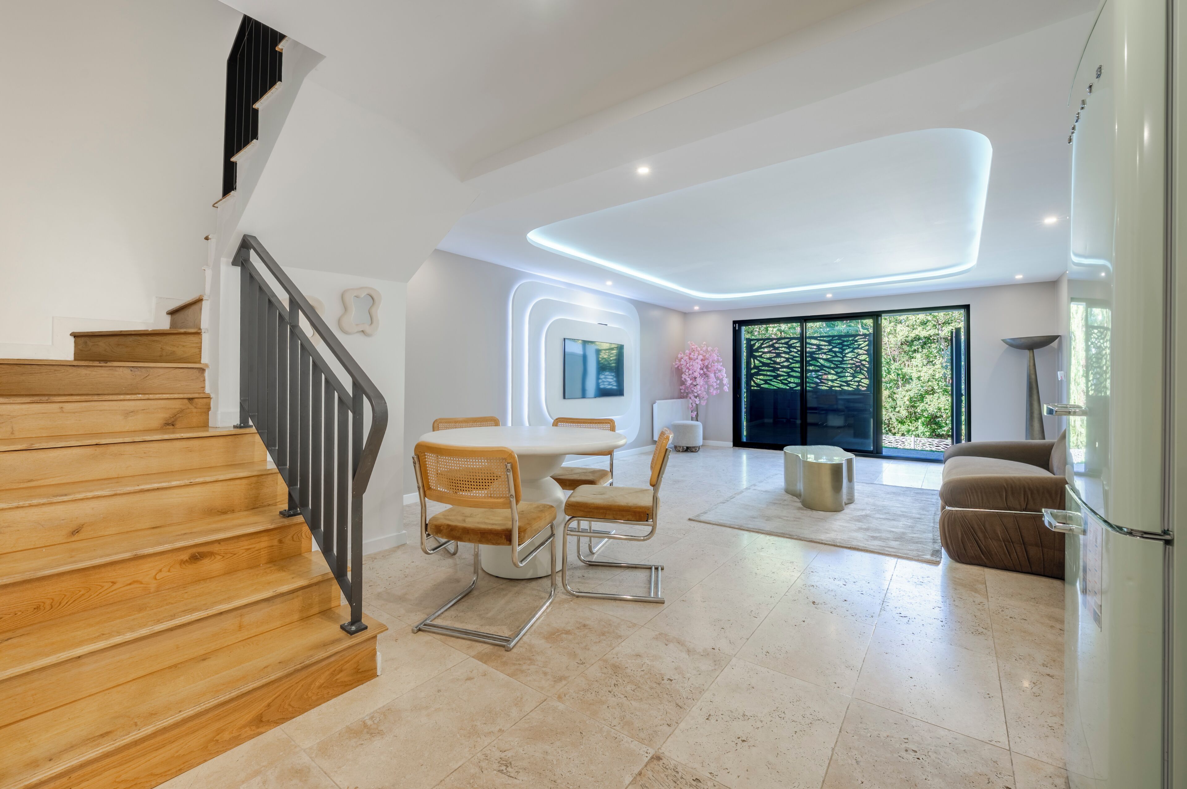 A modern interior with a wooden staircase, a dining area with chairs, a sofa, and a large window with a view.