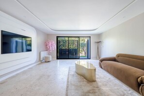 A modern living room with a large flat-screen TV mounted on the wall, a brown sofa, a unique coffee table, and a sliding glass door leading to an outdoor area. - Exceptional Home • 4 Suites • Along the Marne River • Joinville (Joinville-le-Pont)