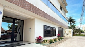 A modern building with a flat roof, large windows, and a paved entrance area with small plants. - ASA Coral Sea Gravata 2BR (Navegantes)