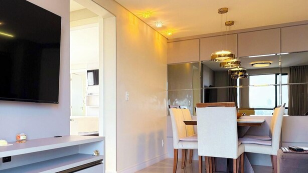A modern living room with a flat-screen TV, a dining area with a table and chairs, and a large window providing natural light. - ASA Coral Sea Gravata 2BR (Navegantes)