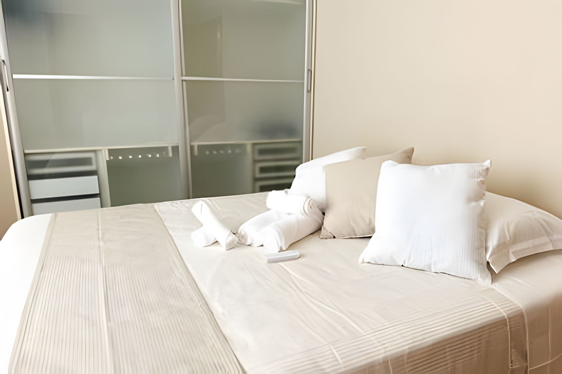 A neatly made bed with white linens and pillows, a remote control, and a window with frosted glass panels.