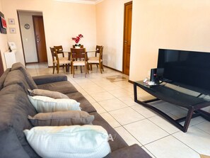 A living room with a sofa, a coffee table, and a TV. There is a dining area with chairs and a table in the background. - ASA Apto Praia Central Amplo 2QT BC (Balneário Camboriú)