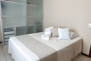 A neatly made bed with white linens and pillows, a glass door closet, and a tiled floor. - ASA Apto Praia Central Amplo 2QT BC (Balneário Camboriú)