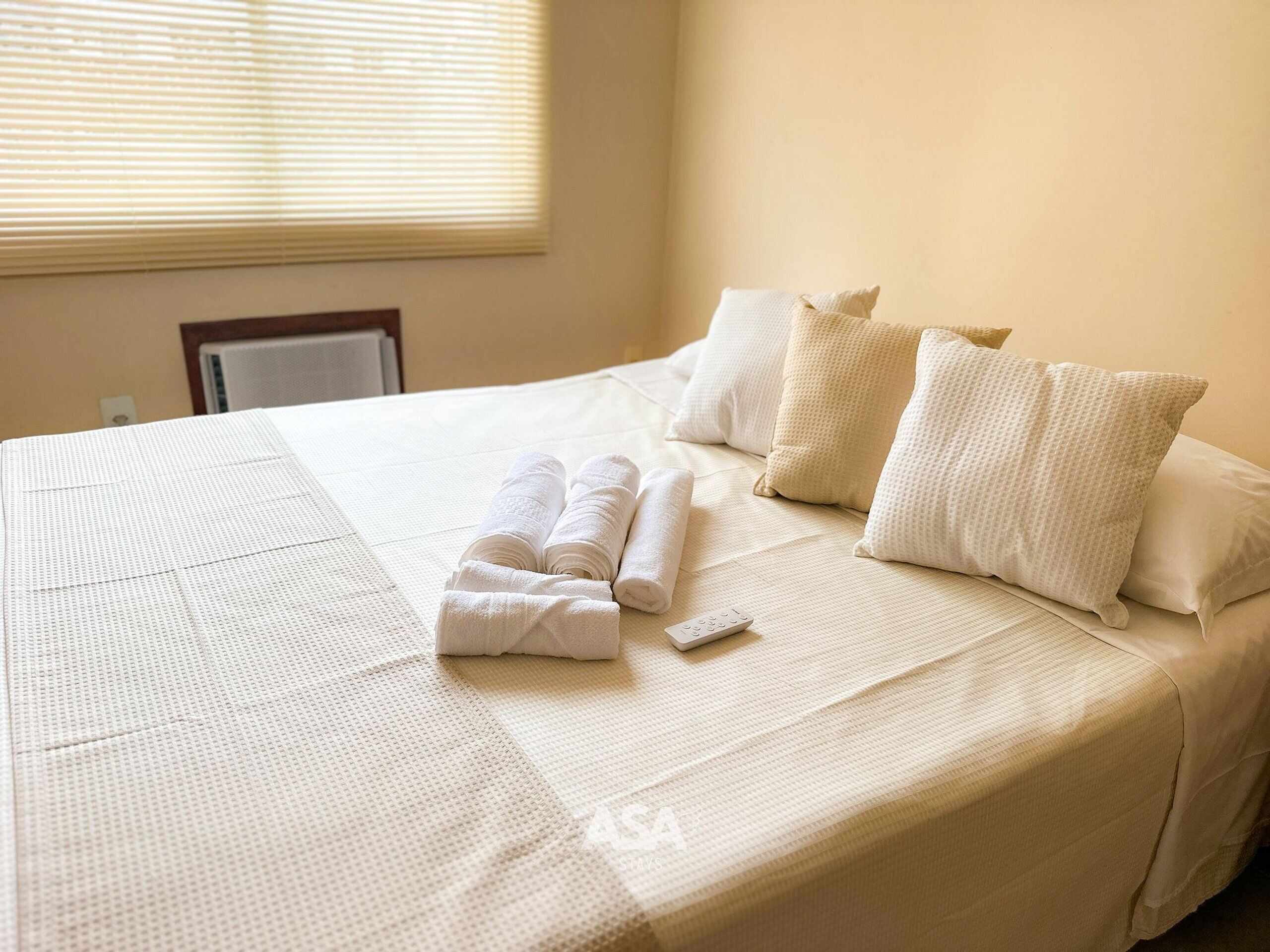 A neatly made bed with rolled towels and a remote control, a window with blinds, and a plain wall.