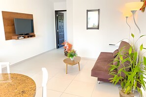 Una sala moderna con un televisor de pantalla plana montado en un panel de madera, un sofá marrón, una mesita redonda con un jarrón de flores y una planta en maceta.