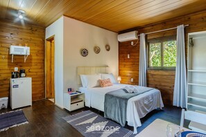 A cozy bedroom with a wooden ceiling, a bed with a white comforter, a small bedside table, a white heater, and a window with curtains. - Charming and Well-equipped Chalet Rel0002 (Imbituba)