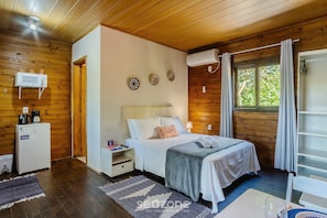 A cozy bedroom with a wooden ceiling, a bed with a white comforter, a small bedside table, a white heater, and a window with curtains. - Charming and Well-equipped Chalet Rel0002 (Imbituba)