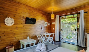 Habitación de cabaña de madera con televisión, una mesita con un farol, un banco y vistas a la vegetación a través de la puerta.