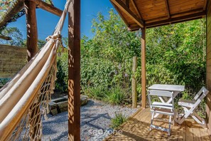 Una hamaca, una mesa de madera y una silla sobre una terraza de madera con un jardín de fondo.
