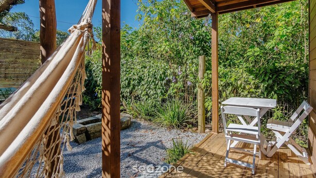 Una hamaca, una mesa de madera y una silla sobre una terraza de madera con un jardín de fondo.